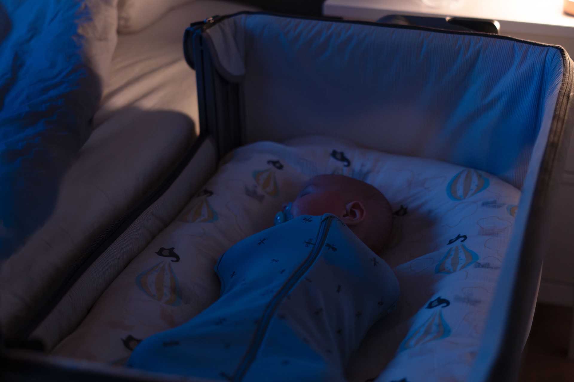 A baby wrapped snugly in cocoon swaddle bag is peacefully sleeping in a crib at night near parent bed. Dimly lit, calm atmosphere ideal for rest. Safety newborn baby sleep. Soft selective focus